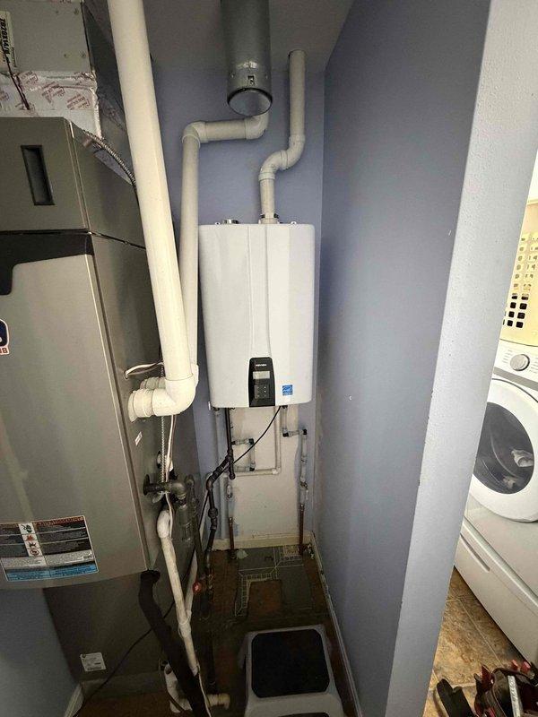 Santa Claus, IN - Installed NPE 240 tankless water heater in residential utility closet, mounting unit to wall and connecting white PVC venting pipes with proper exhaust and intake configuration. Completed all plumbing connections, electrical hookup, and verified unit operation with display panel showing active status, ensuring proper clearances from adjacent HVAC equipment and washing machine in compact mechanical space.